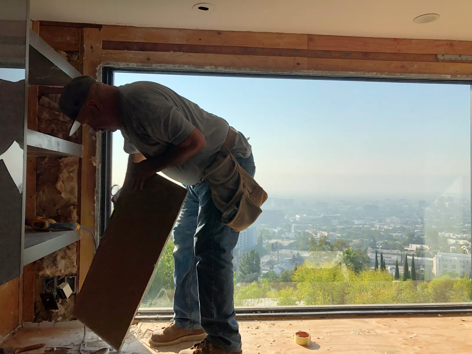 A drywall hanger carrying a sheet of gypsum board into position, silhouetted against a full-height window with the Los Angeles basin behind.
