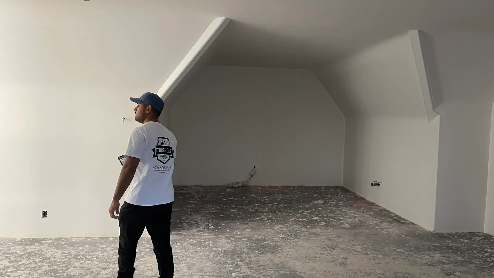 A Stronghold Drywall team member in a branded shirt standing in a finished vaulted room, looking along the wall plane to check the finish.